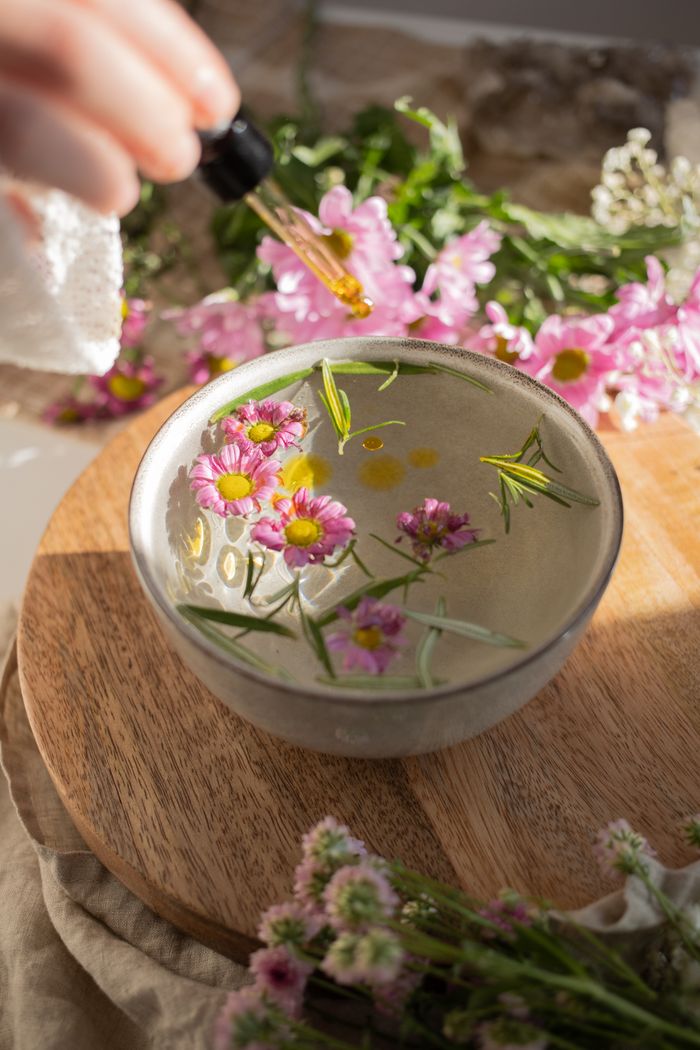 A serene setup of a bowl with floating flowers and essential oil on a wooden board.