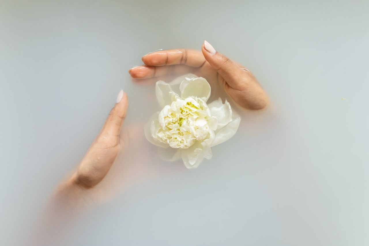 about-img-01 Unrecognizable female with manicured hands holding white flower in hands in soapy water during spa procedures